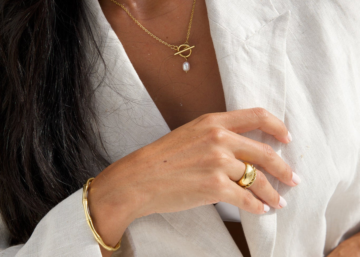 A close-up lifestyle shot of a model wearing a delicate gold necklace with a small white pearl pendant and a matching gold bangle bracelet, styled with a white linen shirt.